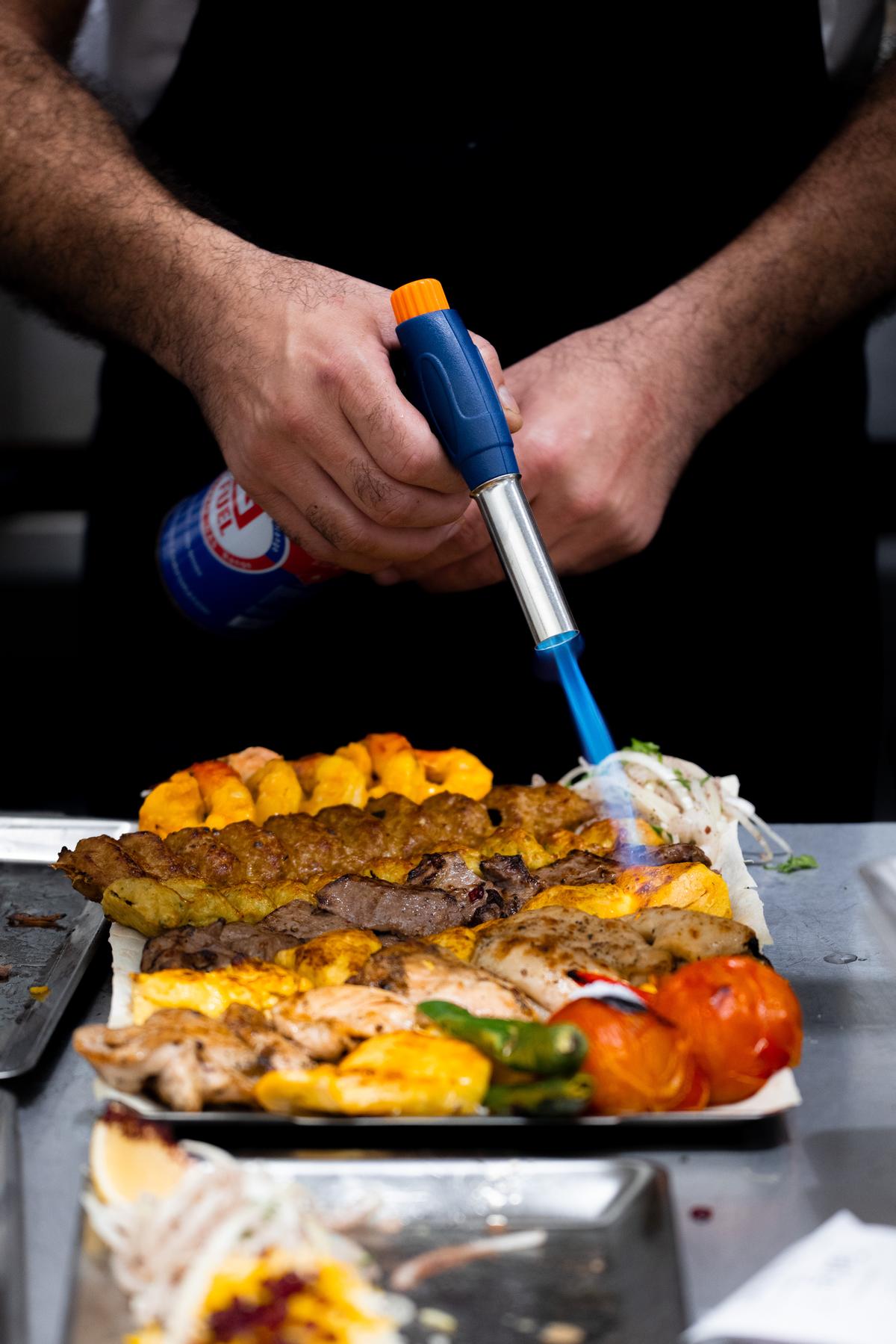 The chef finishing a kebab platter with a torch at the pass